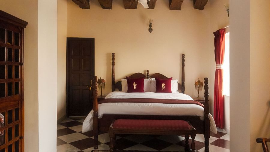 A bedroom at Fort Barli, viewed through an arched doorway, features a four-poster bed, checkered floors, and exposed wooden beams.