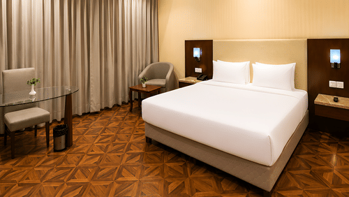 Deluxe room at Golden Tulip Kukas Jaipur with a king bed, white bedding, patterned carpet flooring, grey curtains, and soft ambient lighting.