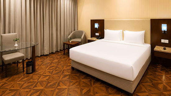 Deluxe room at Golden Tulip Kukas Jaipur with a king bed, white bedding, patterned carpet flooring, grey curtains, and soft ambient lighting.