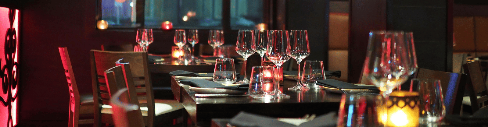 A moody interior shot of a restaurant dining table set with wine glasses and candles, seen through a dark window.