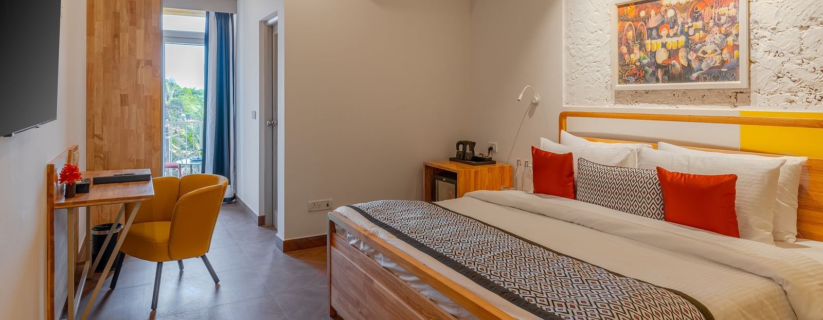 Bright hotel room with, flat TV screen, work desk and chair, vibrant art on the wall, and an access to the balcony at Bedzzz Xclusiv Morjim, Goa