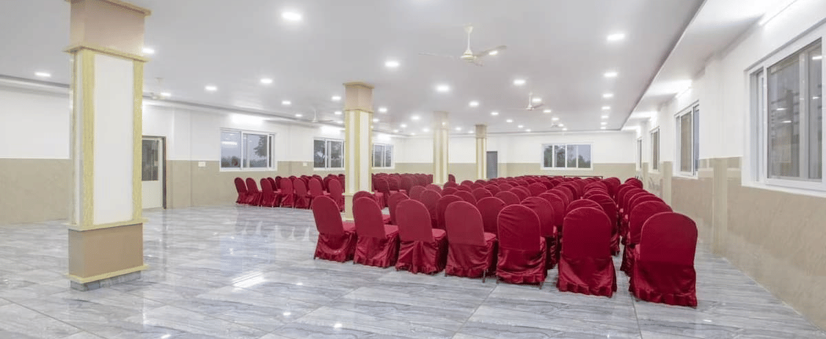 Back view of a spacious banquet hall at Siri Ambari Resorts and Spa, featuring red-covered chairs and bright lighting.