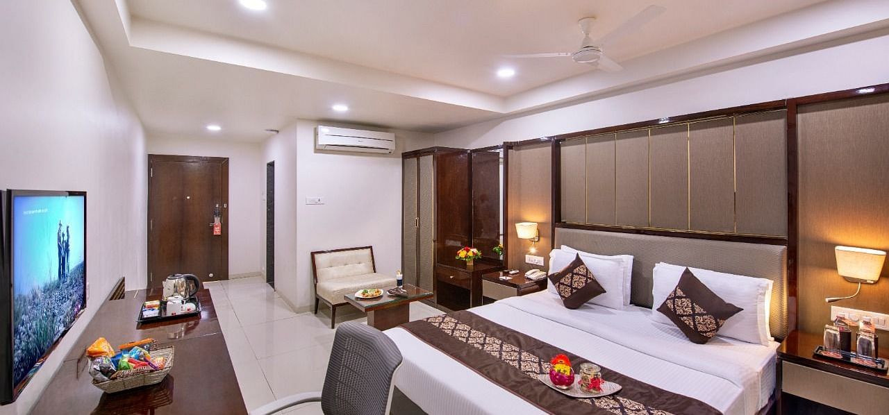 A well-lit hotel room offers a king size bed with decorative pillows, a work desk with a television, and a glimpse into the bathroom.