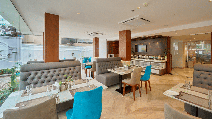 Wide view of the restaurant at QWhite Crescent Luxury Hotel by Dimora Kochi with teal chairs, cushioned booths, and warm ambient lighting.