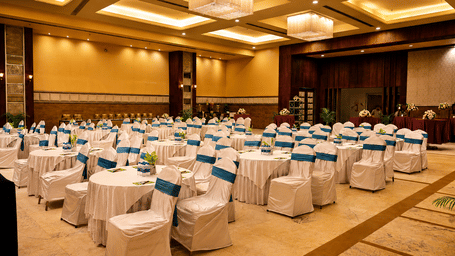 Banquet hall set up theatre-style with rows of white chair covers and teal sashes, chandeliers overhead and warm gold ambient lighting round.
