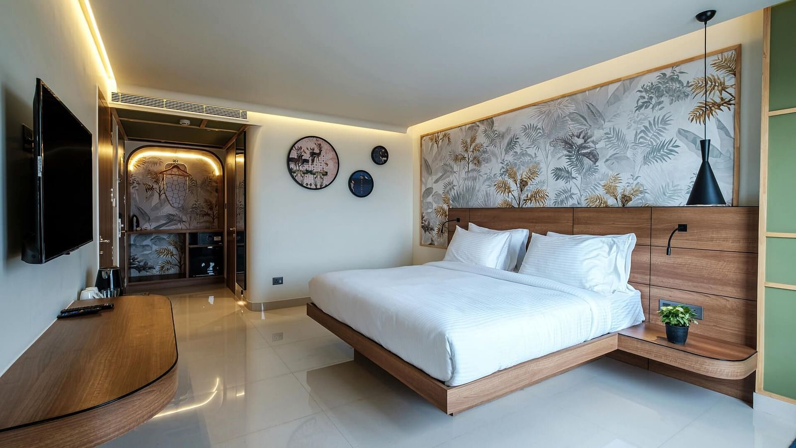 Spacious room at Indie Stays, Jaipur, featuring a large bed with a wooden headboard, a wall-mounted TV, and a pathway to the entrance.