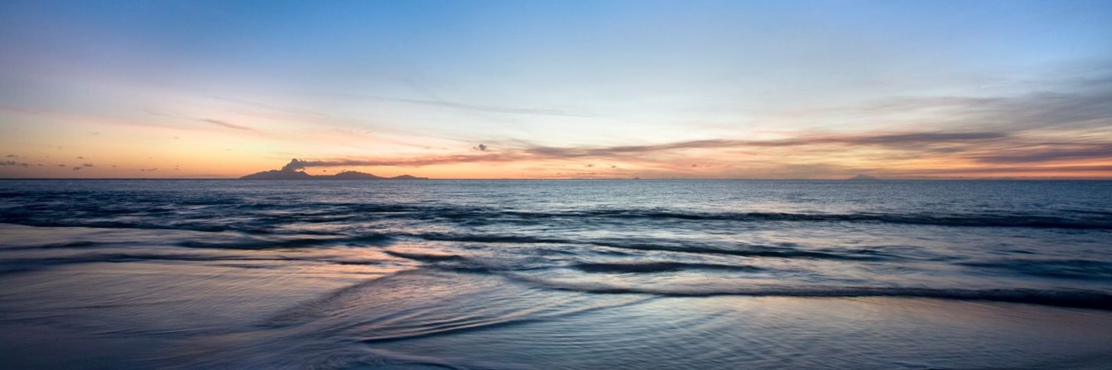 Peaceful Caribbean shoreline at sunset with soft waves, golden hues on the horizon, and a tranquil beach atmosphere.