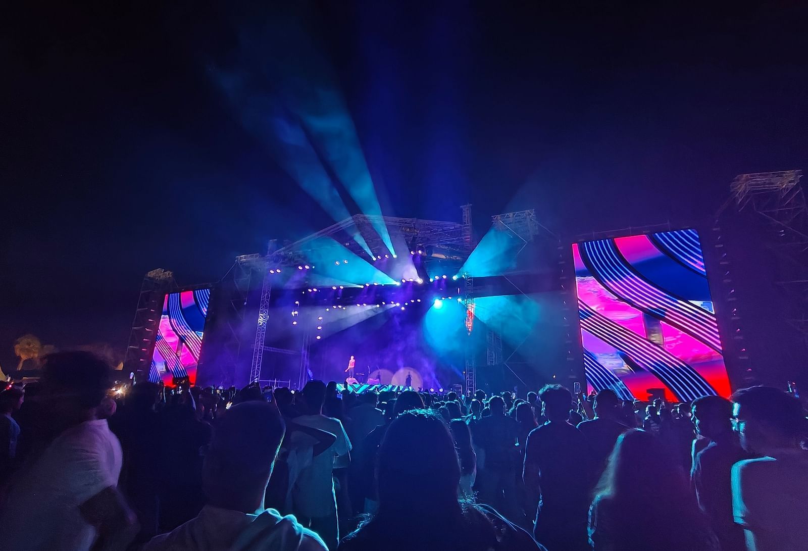 A crowd of silhouetted figures is gathered in front of a stage illuminated by bright lights. The stage features abstract shapes and figures, suggesting a live music performance or event.