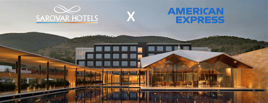 A building with several windows, restaurant with glass walls, and a corridor with plants and lights. A promotion for Amex Card holders at Sarovar Hotels.