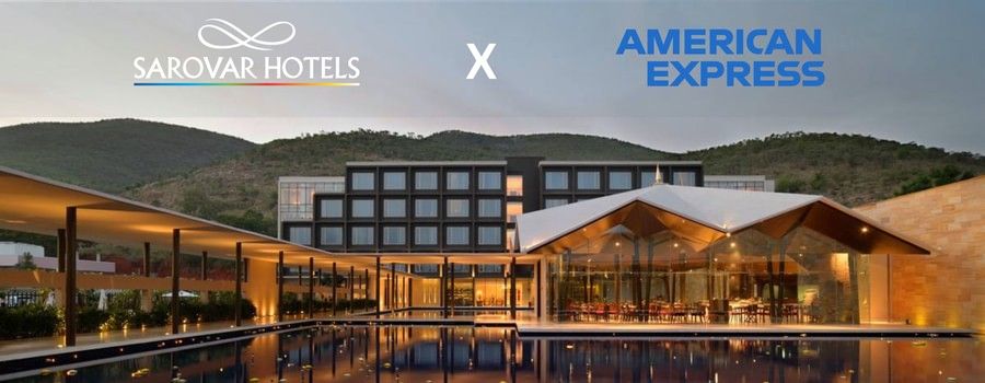 A building with several windows, restaurant with glass walls, and a corridor with plants and lights. A promotion for Amex Card holders at Sarovar Hotels.