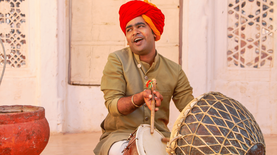 A man in a turban plays a dholak drum while singing indoors at Amantra Shilpi Resort, Udaipur.