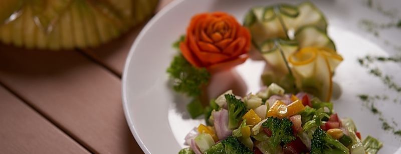 A plate of vegetable salad made with diced cucumbers, tomatoes, onions, and herbs, garnished with a carved tomato rose served on a wooden table at The Hamuse Hotel, Kodaikanal.
