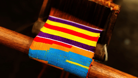 A wooden weaving loom with multicoloured threads being used to create a striped fabric.
