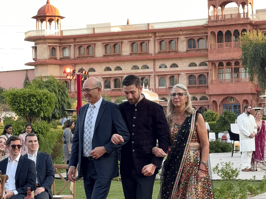 A wedding procession walking down a paved outdoor pathway with the grand, multi-story heritage facade of Umaid Palace visible in the background.