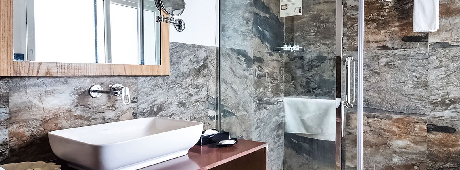 A modern bathroom with a glass shower, a wash basin on a dark counter, and a large mirror.- Bamboo Saa Resort & Spa, Udaipur