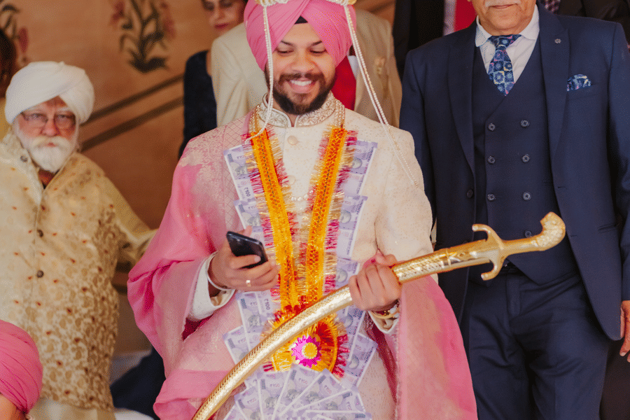 A groom in a pink turban holds a traditional ceremonial sword while standing with guests during an indoor wedding function at Umaid Palace.