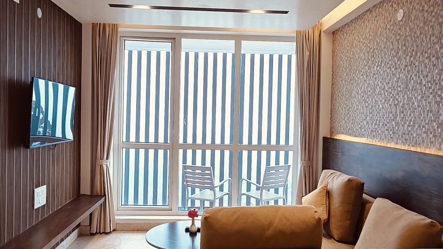 A living room features a beige sofa facing a television on a dark wood wall next to large windows at Bliss Casa Mila By Bergamont Hotels, Kodaikanal.
