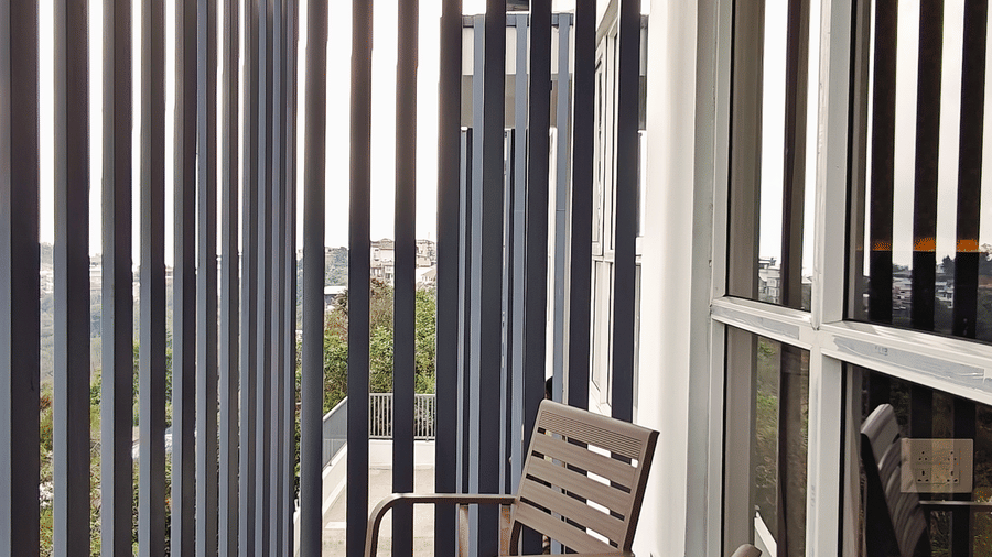 2 chairs are arranged on a long, narrow balcony enclosed by vertical grey slats at Bliss Casa Mila By Bergamont Hotels, Kodaikanal.