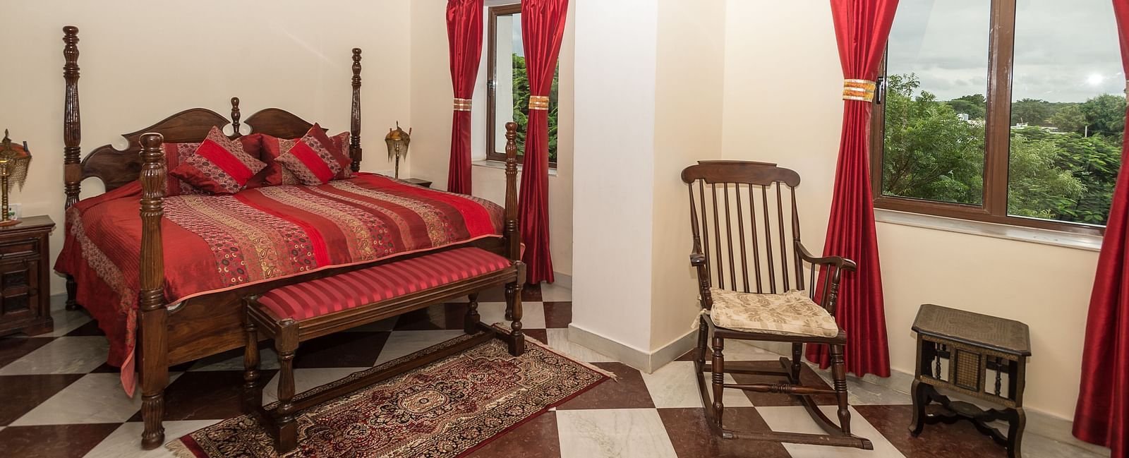 A spacious bedroom at Fort Barli, featuring a four-poster bed with red accents, checkered floors, and exposed wooden beams, offers a blend of rustic charm and elegance.