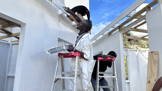 Workers on ladders painting the white exterior trim and beams of a new building structure under a blue sky.