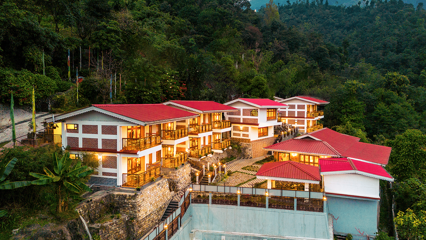 A hillside voyage songfum retreat with multiple buildings featuring red roofs and glowing lights, surrounded by dense green forest and mountains in the background.