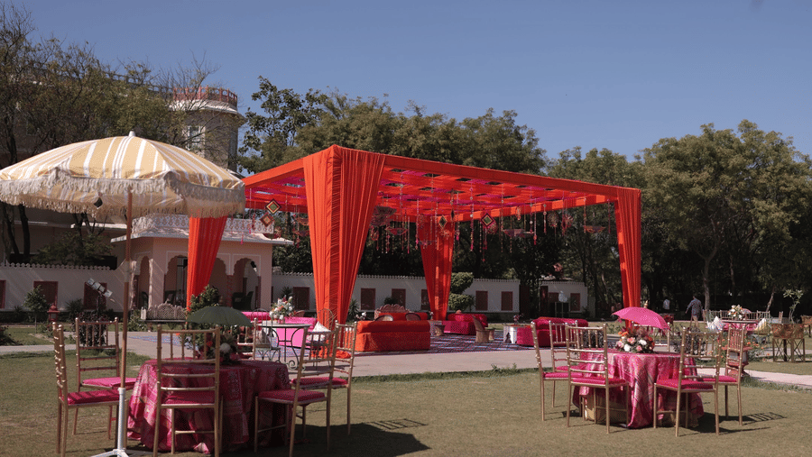 A vibrant outdoor event setup on a lawn featuring a bright orange canopy and pink table arrangements under a clear sky - Umaid Palace