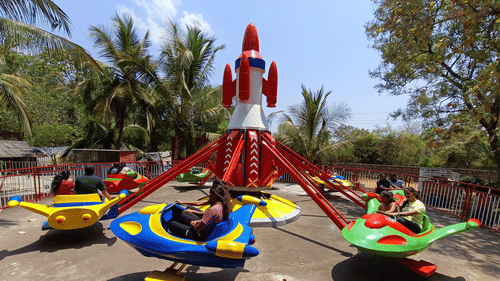 Children and adults enjoying a colourful rocket-themed amusement ride under palm trees and a sunny sky, with a red-and-white rocket centerpiece and spinning, brightly coloured spacecraft seats.