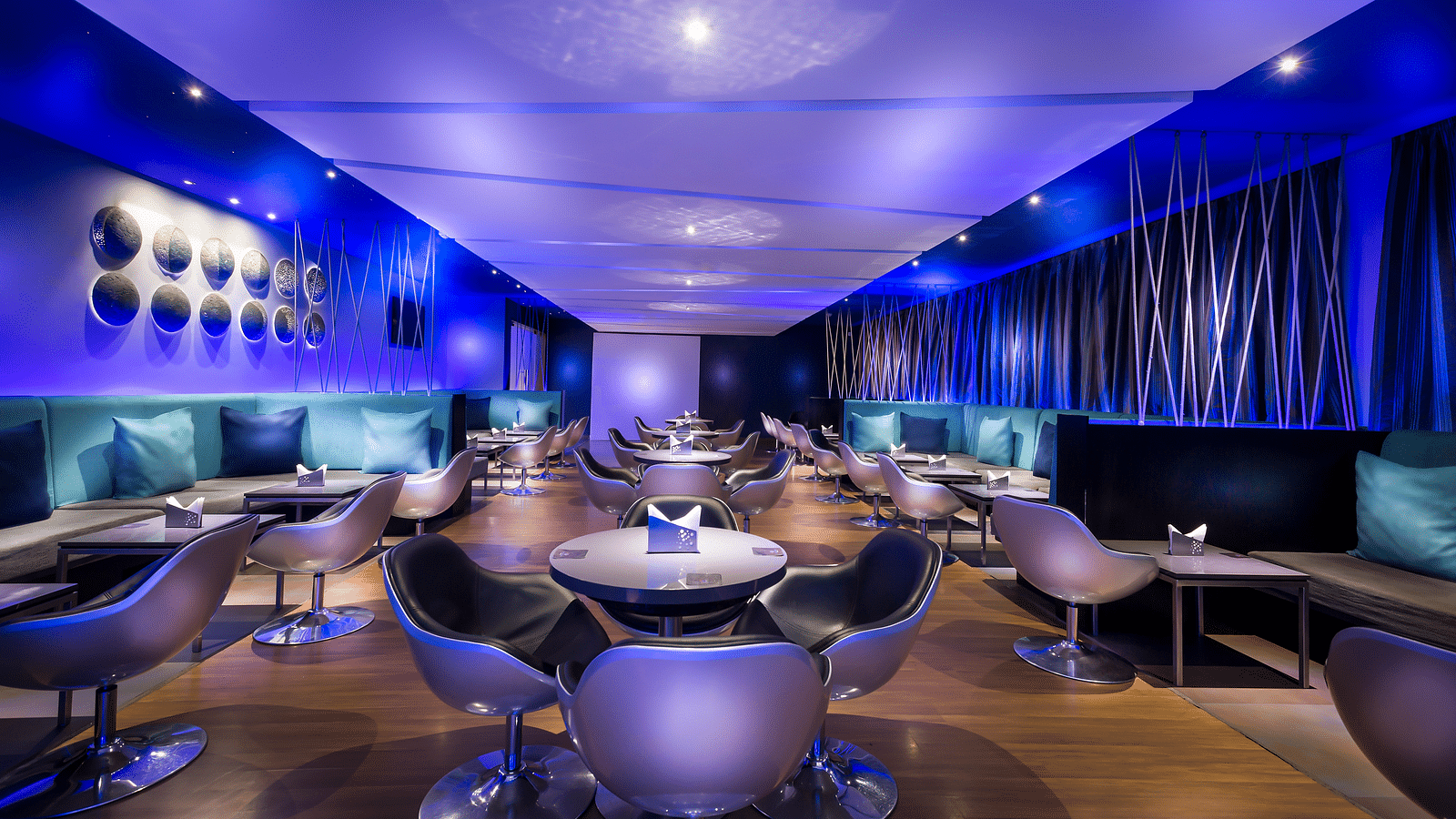 Modern restaurant interior with a blue and purple color scheme. The room features curved seating, round tables, and ambient lighting.