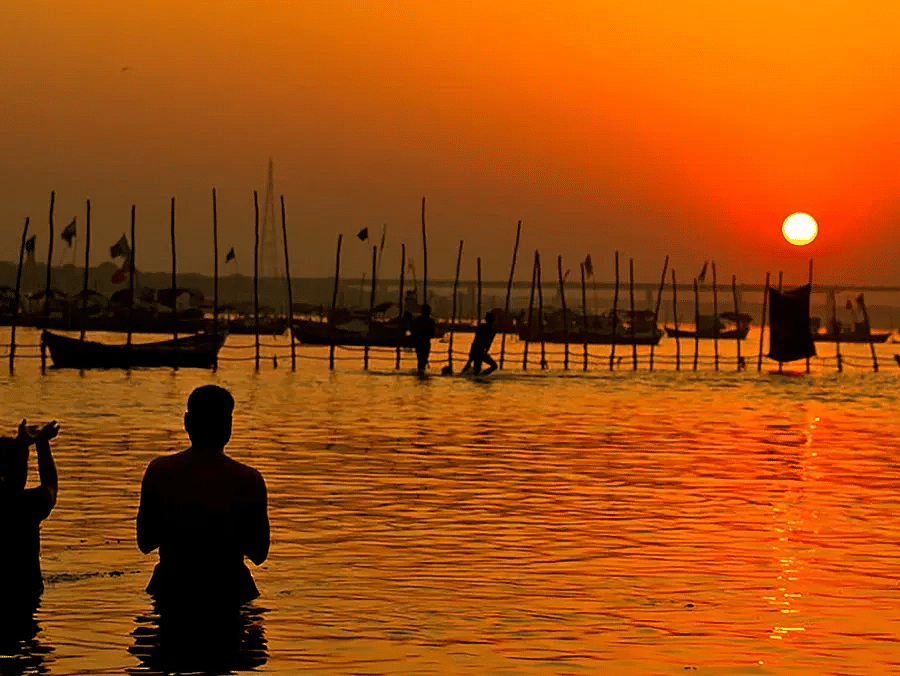 People standing in a river during sunset with boats and structures in the background.