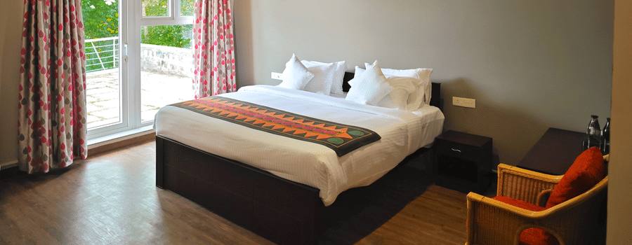 Bedroom in Starling room at Ibex Resorts, Coonoor (Tapas). A bed sits in the middle of the room with an armchair beside it. Curtains cover the windows in the background.