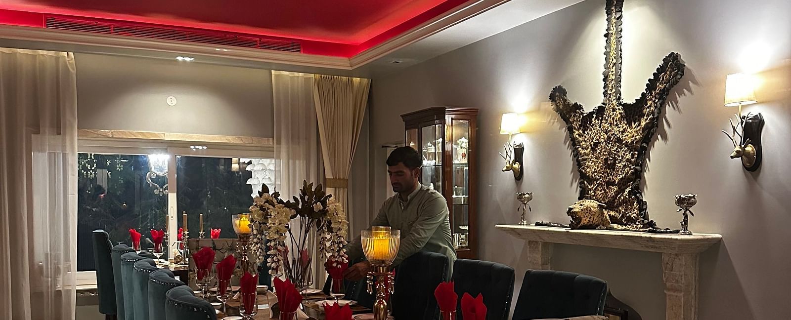 A long dining table set for a meal, with plush chairs, a patterned rug, and a decorative fireplace, under a red ceiling with neon lighting | Dileep Kothi, Jaipur