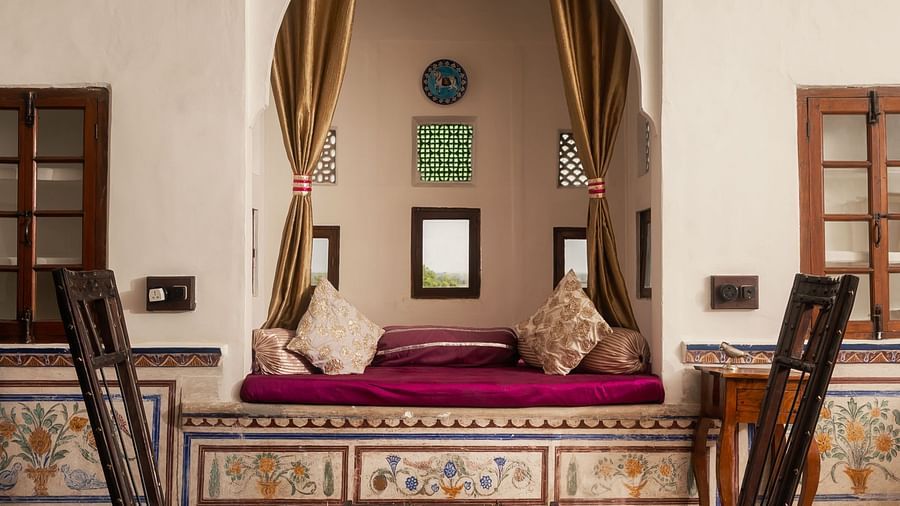 A stylish seating area at Fort Barli, featuring tall chairs, a low table, and an alcove with plush cushions, is illuminated by a striking blue chandelier.