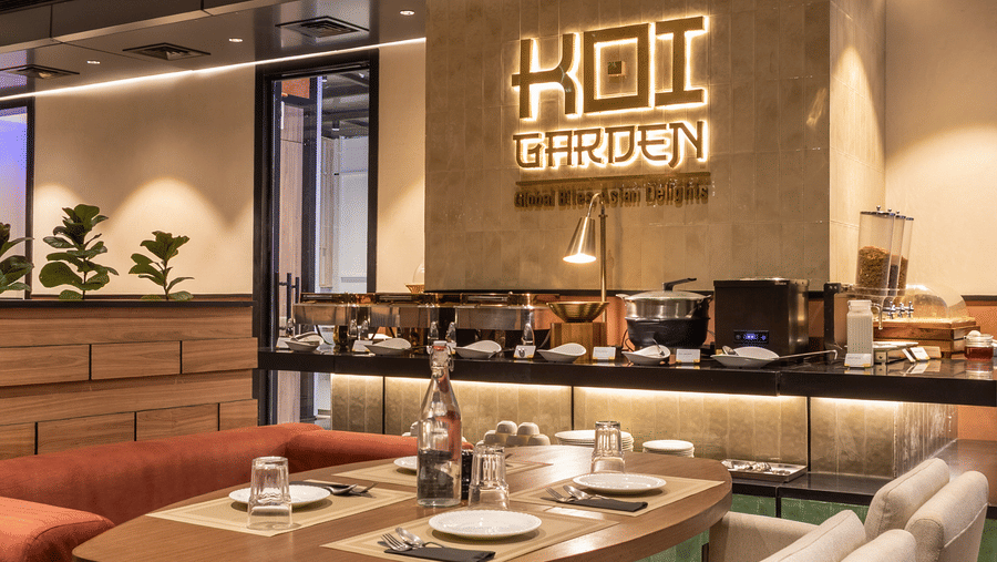 A close-up view of a dining layout inside Koi Garden restaurant. A table is set with plates, cutlery, and glasses between chairs and a booth. The restaurant logo is visible on the background wall.