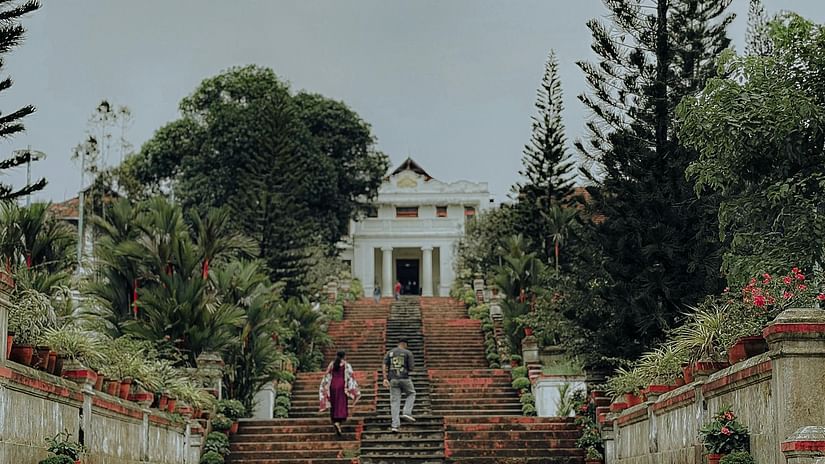 Long staircase flanked by greenery leads to a grand building atop a hill, with cloudy skies overhead.