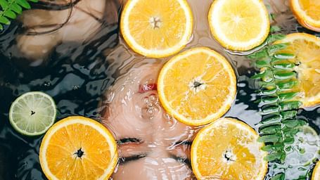 A lady lying submerged in water with her hair spread out, surrounded by fern leaves and orange slices floating on the surface