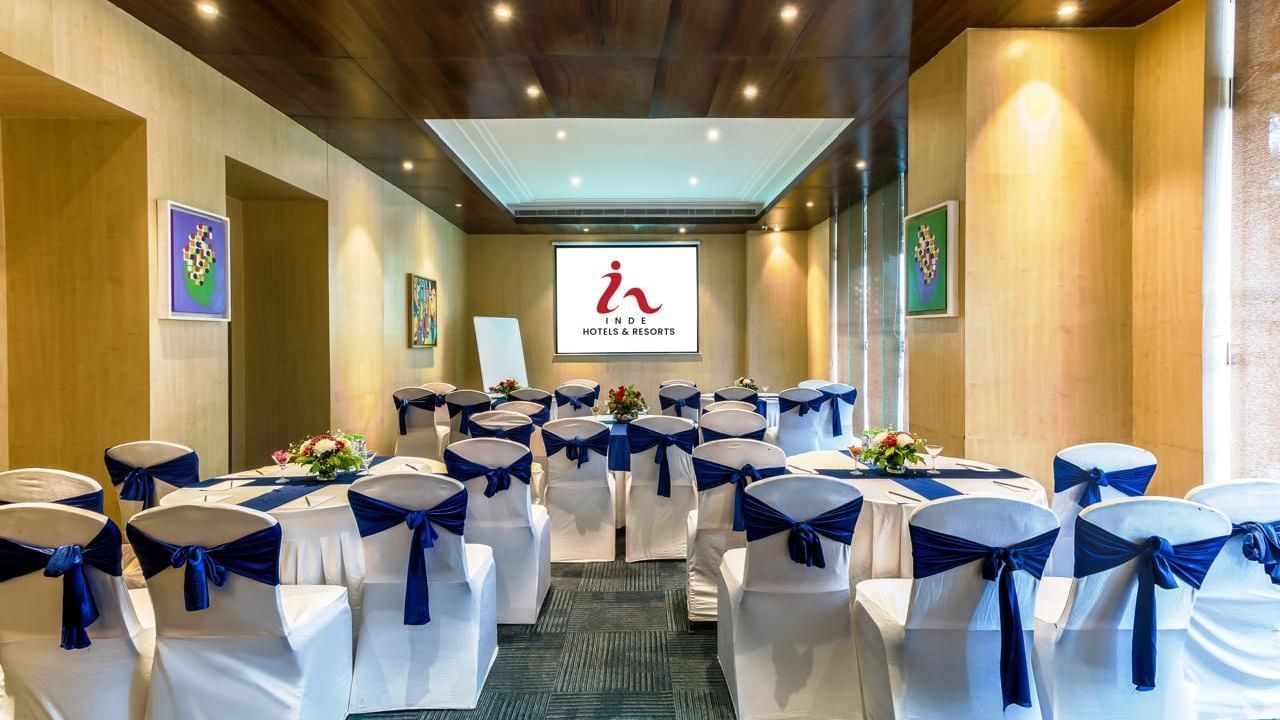 Meeting room at Inde Reva Resorts Rishhikesh, with white chairs tied with navy blue sashes, round tables decorated with flower arrangements, and a projector screen at the front.