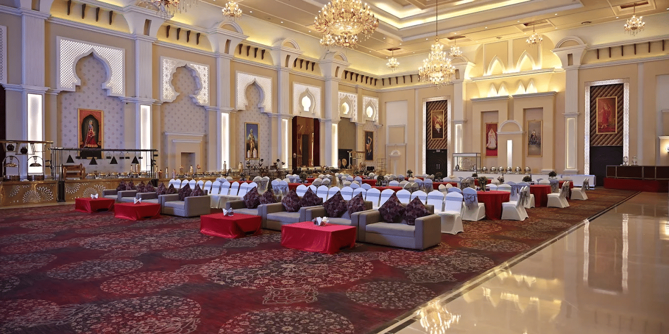 Banquet hall at Geeta Sarovar Portico, Panipat featuring table, chairs, and sofa sets in classroom setting. It also buffet counters, chandeliers handing from the ceiling, and huge paintings on the wall.