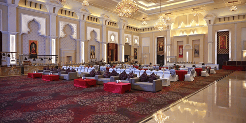 Banquet hall at Geeta Sarovar Portico, Panipat featuring table, chairs, and sofa sets in classroom setting. It also buffet counters, chandeliers handing from the ceiling, and huge paintings on the wall.