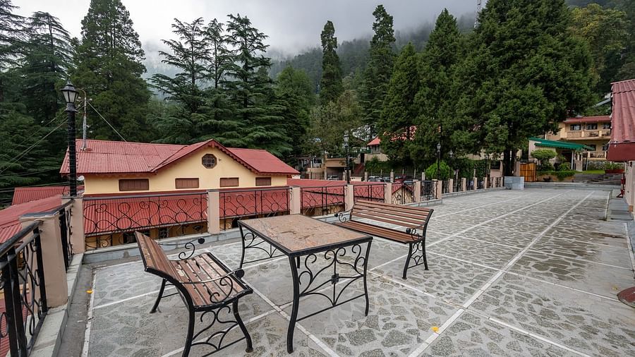 Stone-paved terrace at The Pavilion, Nainital featuring wrought iron benches, mountain vistas, and traditional red-roofed buildings amid dense forest.