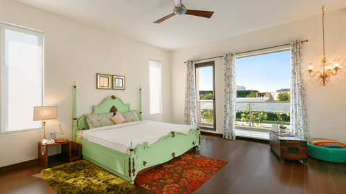 Bedroom with a bed, curtains, wooden floor, large windows, rugs, side table with lamp, chest, cushion, and wall sconce - Karma Lakelands