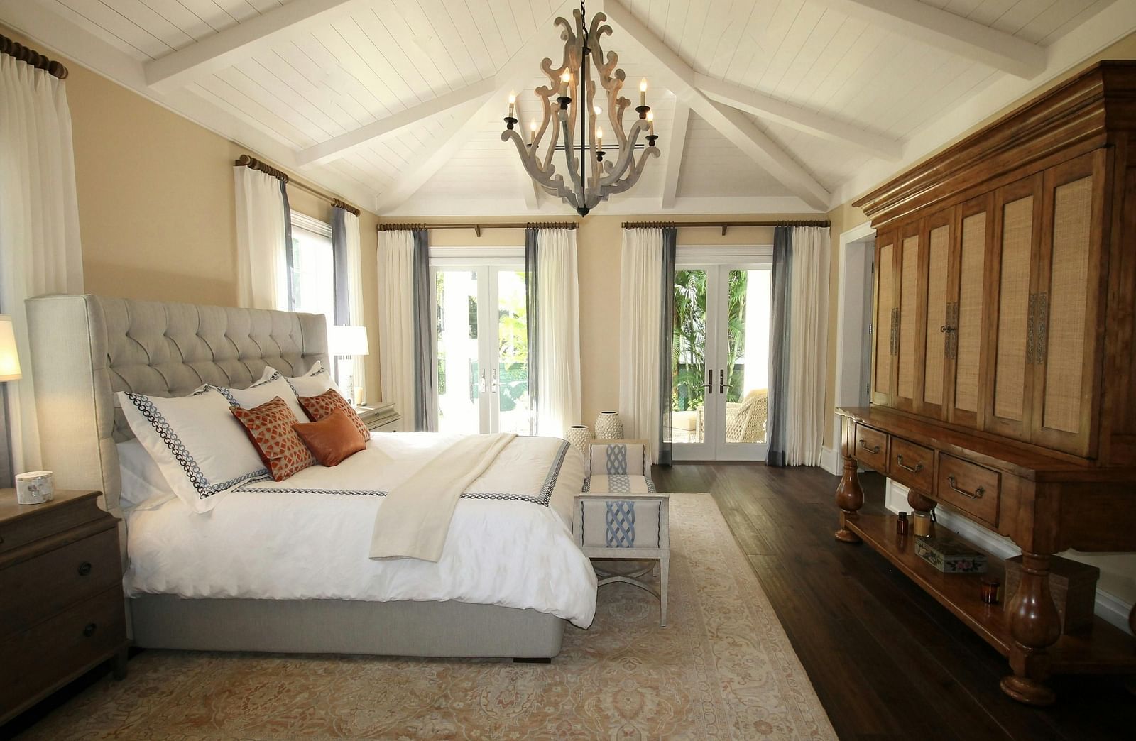 Elegant bedroom with vaulted ceiling, large bed, chandelier, wooden wardrobe, and balcony doors letting in natural light.