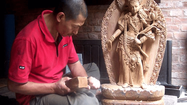 A stone carver working carefully beside a completed traditional Hindu deity statue in a historic masonry workshop in Patan, Nepal.