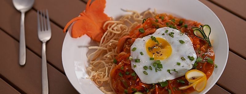  A plate of spaghetti topped with tomato sauce, a sunny-side-up egg, sliced vegetables, and half a boiled egg, served with a fork and spoon on a wooden table at The Hamuse Hotel, Kodaikanal.