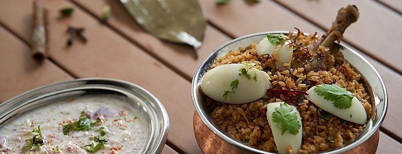 A copper bowl of biryani garnished with boiled egg slices and herbs, accompanied by a bowl of raita at The Hamuse Hotel, Kodaikanal.