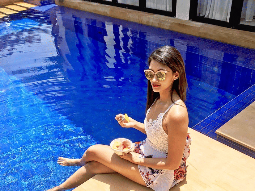 A young woman in a white top sits by the edge of a bright blue swimming pool with her feet in the water.