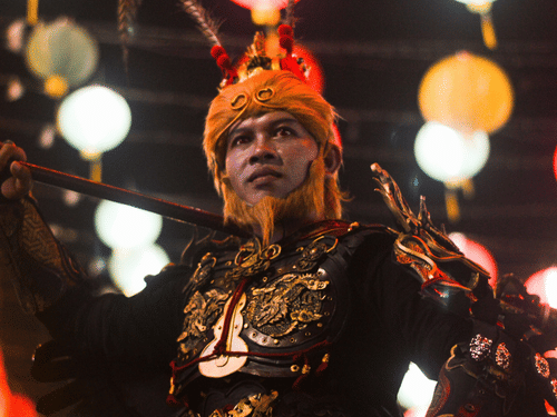 A performer dressed in a  black and gold outfit with a matching headdress, standing amid bright, colorful lights