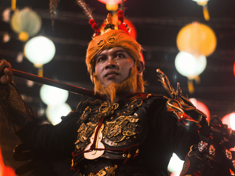 A performer dressed in a  black and gold outfit with a matching headdress, standing amid bright, colorful lights