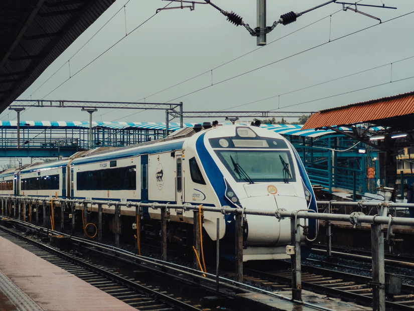 A white and blue Vande Bharat Express train at a railway station platform under overhead electric lines.