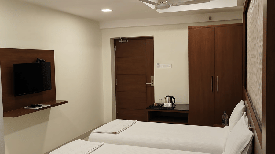 A brightly-lit room, featuring twin beds with white linens, a wall-mounted TV, and dark wood furnishings | A & M Rooms and Residences