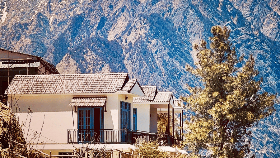 A row of cottages with dark windows is situated on a hillside, overlooking a vast, rugged mountain range under a clear blue sky at the location of Faraway Cottages, Auli.